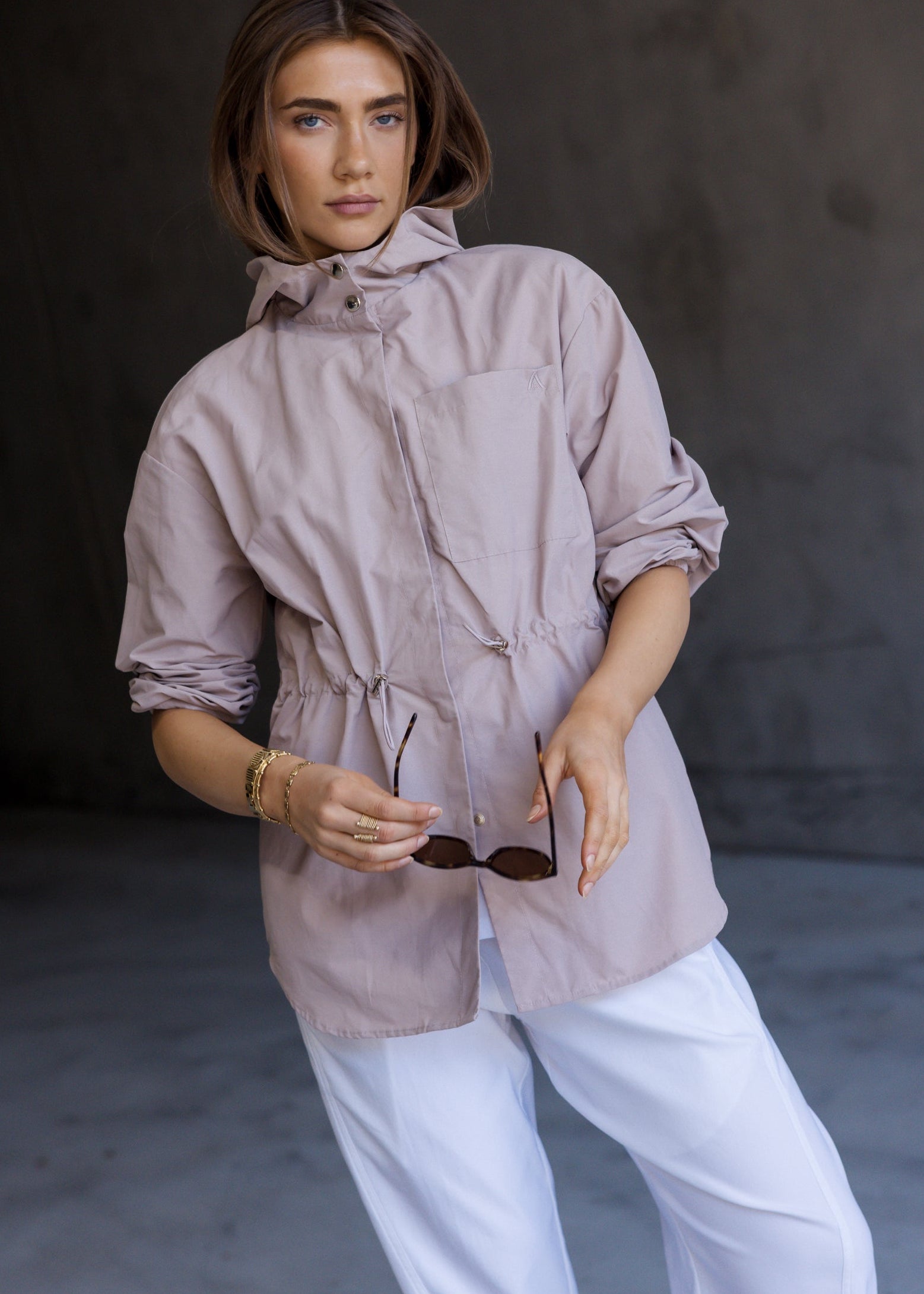 A woman modeling a light taupe hooded jacket with rolled-up sleeves and white pants, holding sunglasses against a dark gray concrete background.