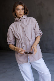 A woman modeling a light taupe hooded jacket with rolled-up sleeves and white pants, holding sunglasses against a dark gray concrete background.